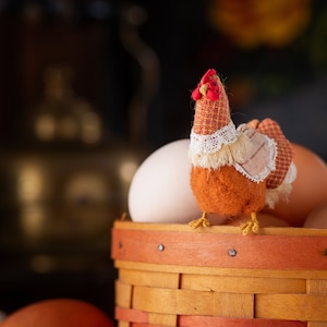 Puede incluir: Una figurita de pollo de peluche marrón y blanco se sienta encima de una cesta de huevos marrones. La cesta está hecha de madera tejida con acentos rojos.