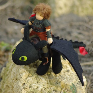 May include: A handmade felt doll with brown hair riding a black felt dragon with green eyes. The dragon has wings and a red tail. The doll is wearing a brown leather vest and pants with a black shirt. The dragon and doll are sitting on a rock.