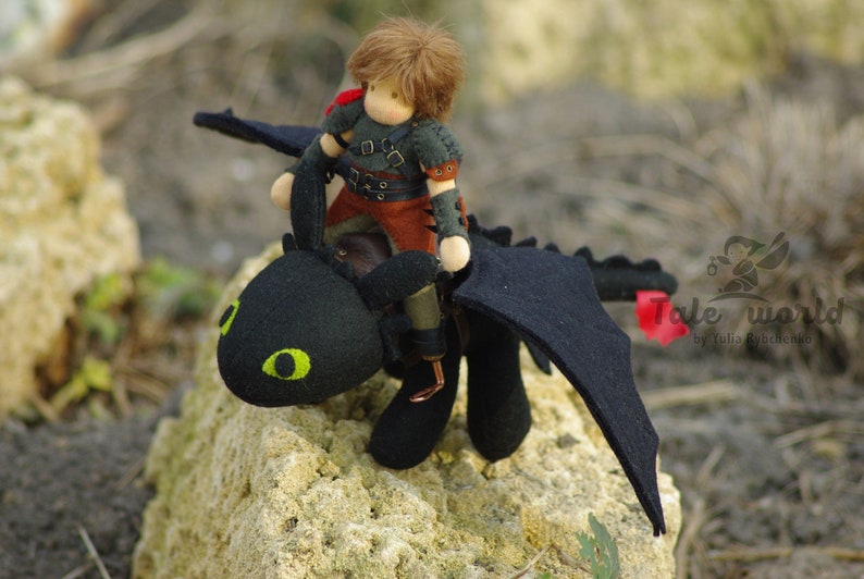 May include: A handmade felt doll with brown hair riding a black felt dragon with green eyes. The dragon has wings and a red tail. The doll is wearing a brown leather vest and pants with a black shirt. The dragon and doll are sitting on a rock.