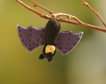 Könnte beinhalten: Eine lila Filzfledermaus mit schwarzem Körper und einem gestickten Gesicht hängt an einem Ast. Die Fledermaus hat ein gesticktes Lächeln und Augen.