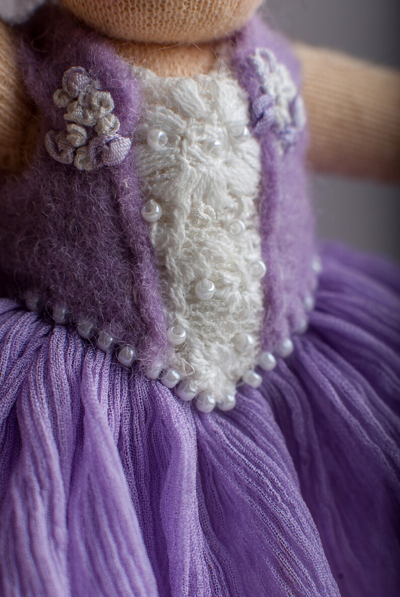 May include: A purple knitted doll dress with a white lace bodice and pearl embellishments.