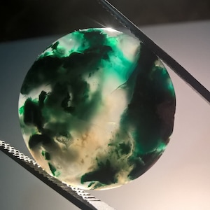 May include: A round, green and white agate cabochon with a swirling pattern. The stone is held in tweezers.