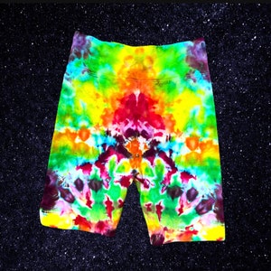 May include: Vibrant tie-dye biker shorts in a spectrum of colors including yellow, red, green, and purple. The shorts have a high waist and a fitted design. The background is a dark, starry space.