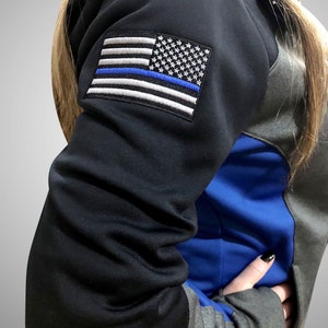 May include: Black and gray hooded sweatshirt with a blue accent and a patch of the American flag with a blue stripe. The sweatshirt has a front pocket and a black sleeve.