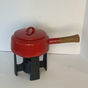 May include: Red enamel fondue pot with a wooden handle and a black metal stand. The pot has a lid with a small handle.