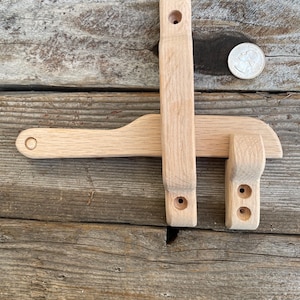 Könnte beinhalten: Ein hölzerner Torriegel, bestehend aus einem horizontalen Riegel und einem vertikalen Teil. Der Riegel ist aus hellem Holz gefertigt und dient zur Sicherung eines Tores oder einer Tür. Eine Münze liegt neben dem Riegel. Der Riegel ist etwa 20 cm lang.