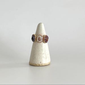 May include: A gold ring featuring three decorative circular stones. The stones are in red, white, and black, set in a gold band. The ring is displayed on a white ceramic cone.