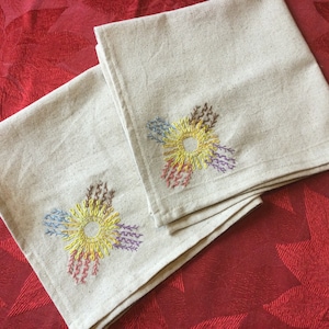 May include: Two beige linen kitchen towels with embroidered colorful flowers in the corner. The flowers are a multi-colored sunburst design.