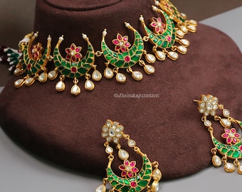 May include: A gold-coloured necklace and matching earrings set. The necklace and earrings feature green and red enamel crescent-shaped designs with floral accents and dangling teardrop-shaped white gems. The jewellery is displayed on a brown mannequin head.