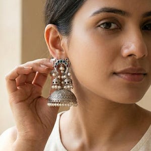 May include: Silver-coloured, ornate earrings with a bell-shaped design. The earrings feature intricate patterns, small pearls, and colourful accents. The earrings are being held up to the ear, showcasing their design and details.