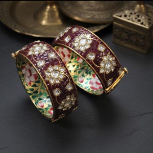 May include: Two ornate, burgundy bangles with floral and diamond-like accents. The interior of each bangle features a colourful floral pattern. Gold-coloured metal is used for the clasp and trim. The bangles are displayed on a dark surface.