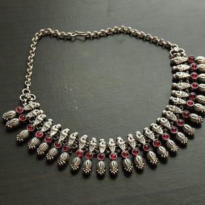 May include: Silver-toned necklace with a chain and ornate pendant design. The necklace features small, round, red gemstones set within the pendants. The necklace is displayed on a dark surface.
