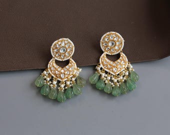 May include: A pair of gold-toned earrings with a circular top and a crescent-shaped bottom, adorned with white stones and small pearls. The earrings feature teardrop-shaped green stones that dangle from the bottom.