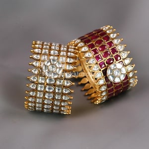 May include: Two gold-toned bangles with intricate designs. One features clear, square-cut stones and a central circular design. The other bangle has red stones and a similar circular design. Both are set against a gray background.