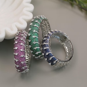 May include: Three silver bangles with a geometric design, each set with small stones in different colors: green, purple, and blue.