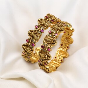 May include: Two ornate gold bangles with intricate designs and embedded red and clear gemstones. The bangles are displayed on a white, silky fabric, showcasing their detailed craftsmanship and rich color.