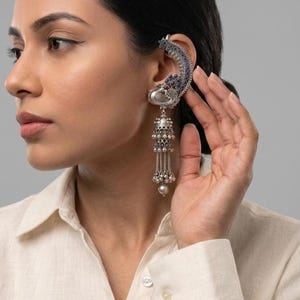 May include: A silver ear cuff with a peacock design, featuring blue accents and dangling beaded elements. The cuff is worn on the ear, showcasing intricate details and a dangling pearl at the bottom. The jewelry piece is a statement accessory.