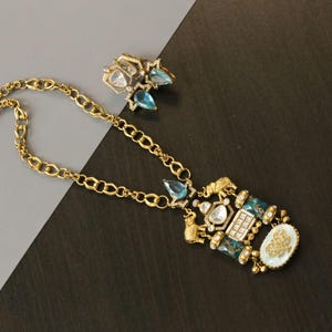 May include: A gold-toned necklace featuring a thick chain and a large pendant with blue teardrop stones, clear crystals, and two small gold elephant figures. The pendant includes a white oval accent with a gold emblem and matching stud earrings.