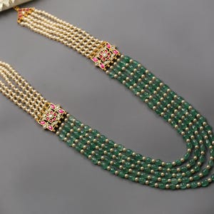 Puede incluir: Un collar de múltiples hilos con hilos alternos de cuentas verdes y pequeñas cuentas redondas de color blanquecino. El collar tiene conectores dorados con detalles en rojo y blanco. El collar se muestra sobre una superficie gris.