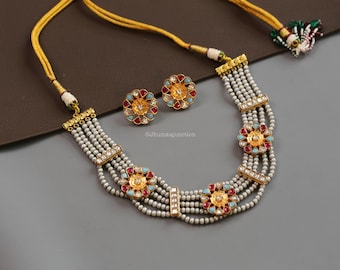 May include: A gold-toned necklace and matching earrings set. The necklace features multiple strands of white beads, accented with gold-coloured floral designs with red and turquoise details. The earrings mirror the floral design.