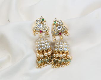 May include: A pair of gold-toned earrings with a peacock design. The earrings feature clear, faceted stones, small pearls, and green beads. The peacock's head has a green and red accent. The earrings hang from a gold chain.