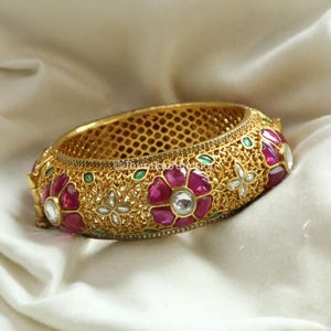 May include: A gold-toned metal bangle bracelet featuring an intricate filigree pattern, embellished with vibrant pink floral-shaped stones, small green leaf-shaped accents, and clear, sparkling white stones set in a repeating decorative design.