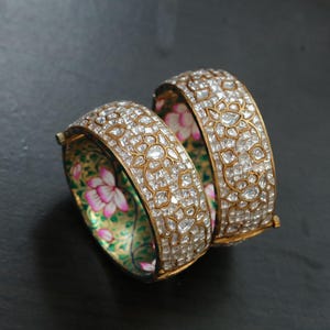 Pode incluir: Dois braceletes articulados em tom dourado com a superfície exterior densamente incrustada de pedras transparentes facetadas num padrão floral. O interior de cada bracelete é decorado com um design pintado à mão de flores de lótus cor-de-rosa.