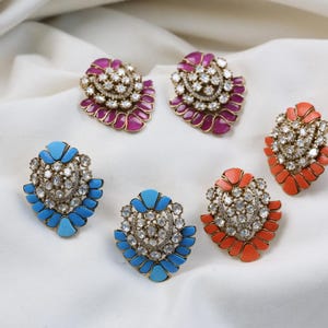 Op de afbeelding: Zes sierlijke oorbellen met juwelen in magenta, blauw en koraal. Elk heeft een centrale cluster van heldere stenen, omgeven door een bloembladachtig ontwerp. Gepresenteerd op een witte, gedrapeerde stof.