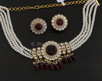 May include: A five-strand pearl choker necklace with a large, ornate pendant featuring a red stone surrounded by clear stones. The pendant has a floral design and is accented with dangling red stones. The necklace comes with a pair of matching earrings with red stones.