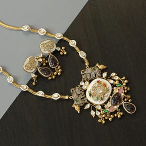 May include: A gold-toned necklace and earring set featuring white enamel accents, elephant motifs, and multi-colored gemstone inlays. The necklace includes a large central pendant with dangling beads, while the matching earrings mirror the design.