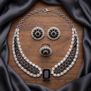 May include: A silver-toned jewelry set featuring a necklace, earrings, and a ring. The necklace and ring have a large black rectangular stone and black and white stones. The earrings are round with black and white stones. The set is displayed on a wooden surface with a gray satin cloth.