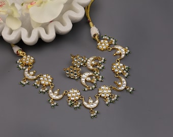 May include: Gold-toned necklace and earring set featuring a crescent moon and floral design. The jewelry is adorned with white pearls and small green beads. The set is displayed on a gray surface, with a white floral accent in the background.