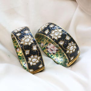 May include: Two hinged bangles featuring a dark navy blue exterior with a textured grid pattern and inlaid white floral motifs. The interior of each bangle displays a hand-painted floral design in green, pink, and white on a gold-toned metal base.