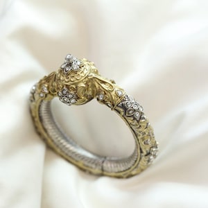 Può includere: Bracciale dorato e argentato ornato con intricati motivi floreali e geometrici. Il bracciale è impreziosito da piccole gemme scintillanti. Il design presenta una chiusura decorativa dettagliata.