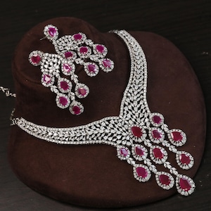 Op de afbeelding: Een schitterende sieradenset met een ketting en bijpassende oorbellen. De ketting en oorbellen zijn versierd met sprankelende heldere stenen en levendige, druppelvormige rode edelstenen. De set wordt gepresenteerd op een bruin fluwelen oppervlak.