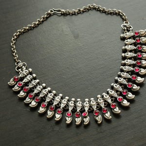 May include: A silver necklace with a chain and a pendant made of multiple teardrop-shaped charms. Each charm has a small, red gemstone in the center.