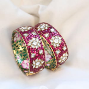 May include: Two ornate bangles with a vibrant design. The bangles feature a deep red background adorned with white floral patterns and accents. The inner surface displays a green and pink floral motif, set against a gold band.