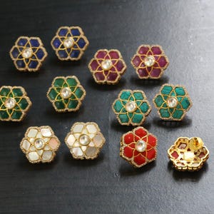 May include: A selection of floral stud earrings in shades of blue, green, red, and white. Each earring has a central stone and a gold-coloured border. The earrings are presented on a dark surface.