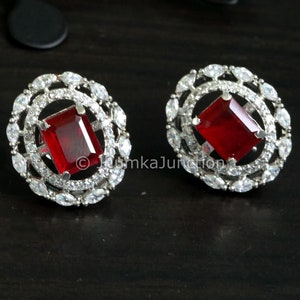 May include: A pair of silver-toned stud earrings. Each earring features a rectangular, deep red gemstone at the center, surrounded by a halo of smaller, clear, marquise-cut stones. The earrings are set against a dark background.