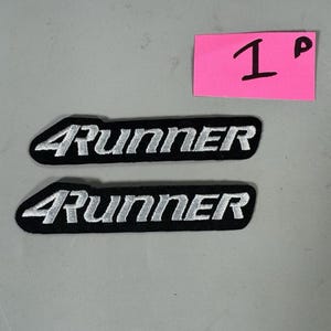 Könnte beinhalten: Zwei schwarze, gestickte Aufnäher mit dem Text "4Runner" in Weiß. Die Aufnäher sind rechteckig mit gewölbter Ober- und Unterseite. Ein pinkfarbener Haftzettel mit der Zahl "1" befindet sich in der oberen rechten Ecke.