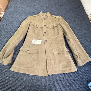 May include: A tan wool-blend military-style jacket featuring a high collar, long sleeves, and four front pockets with buttoned flaps. The jacket has a central button closure and a small white tag attached to the front with the text ER26 written on it.