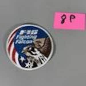 May include: A circular patch featuring a stylized illustration of a falcon with the text "F-16 Fighting Falcon." The patch has a blue background with white stars and red and white stripes, and a brown and white falcon.