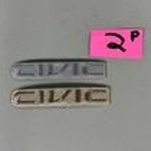 May include: Two silver and gold "CIVIC" car emblems. The emblems are rectangular with the word "CIVIC" in a sans-serif font. The background is a light gray.
