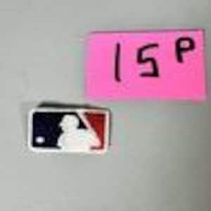 May include: A white, blue, and red MLB logo pin. The pin features the silhouette of a batter in a baseball uniform. A pink sticky note with the number "15" and the letter "P" is next to the pin.