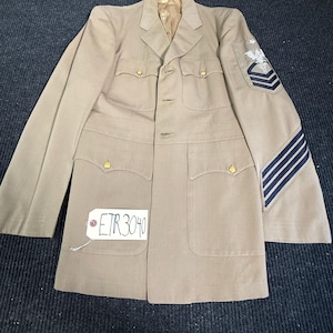 May include: A tan military-style jacket with a notched collar, four front pockets with gold-tone buttons, and a white tag reading ETR 3040. The left sleeve features a white embroidered eagle and caduceus patch above three dark blue chevron stripes.