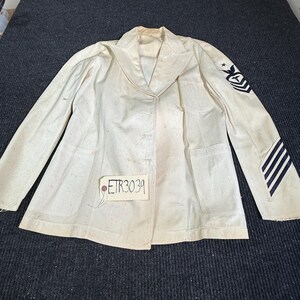 May include: An off-white military-style jacket featuring a notched lapel, long sleeves, and two front pockets. The left sleeve displays a dark blue embroidered rank insignia with a star and stripes. A small tag attached to the front reads ETR3039.