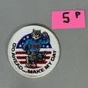 May include: A circular white patch with a cartoon wolf in a blue shirt and red and white striped trousers. The wolf is flexing its arm. The patch has the text "GO AHEAD - MAKE MY DAY" and a pink price tag that says "5 P".