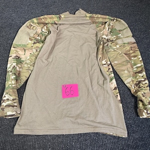 May include: A long-sleeve combat shirt featuring a solid tan, textured torso panel and camouflage patterned sleeves. The sleeves include hook-and-loop patches on the shoulders. A bright pink square tag with the handwritten text 66 is attached.