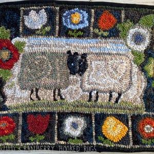 Könnte beinhalten: Handgeknüpfter Wollteppich mit zwei Schafen auf einer Wiese, umgeben von bunten Blumen. Der Teppich hat einen dunkelblauen Hintergrund mit einem schwarzen Rand. Die Blumen sind rot, gelb und blau. Der Text "WILD CRANBERRY HOOKED RUGS" befindet sich unten.