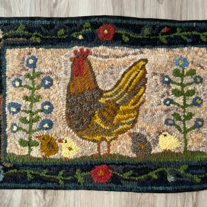 Puede incluir: Alfombra hecha a mano con un gallo y polluelos, rodeada por un borde floral. El gallo es marrón, amarillo y rojo. La alfombra tiene un fondo neutro con flores azules, rojas y amarillas. El borde es azul oscuro.
